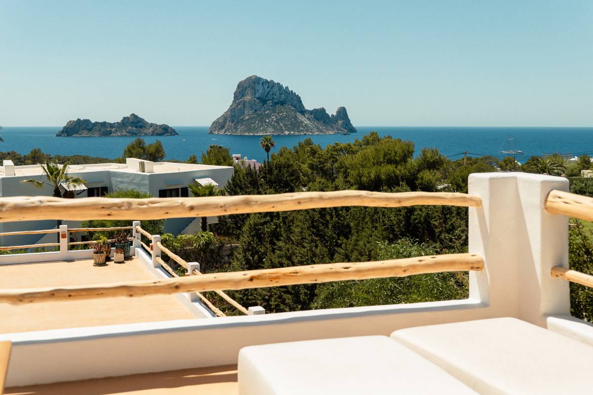 Terrasse ensoleillée avec rambardes en bois surplombant verdure luxuriante, mer bleue et îles rocheuses sous ciel clair.