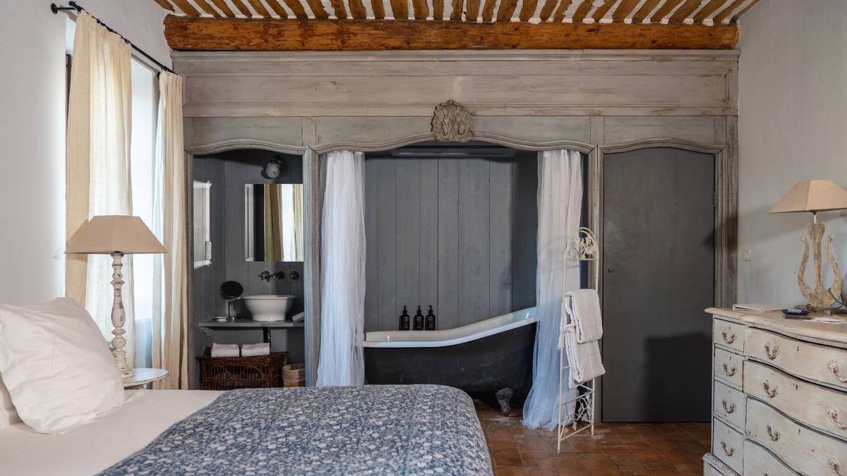 A tidy bedroom features a cozy bed, side table with lamp, shelves, and visible bathtub.