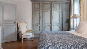A tidy bedroom with cozy bed, side table lamp, and chair near grey shelves.