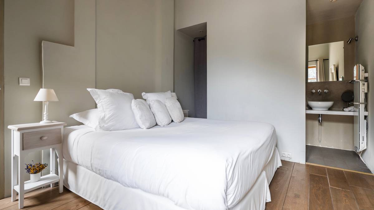 A tidy bedroom features a cozy bed, side table with lamp, and visible bathroom mirror.
