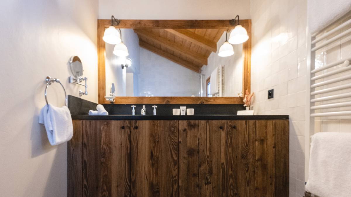  Rustic bathroom with wooden vanity, large mirror, soft lighting, and white tiles under a sloped wooden ceiling.