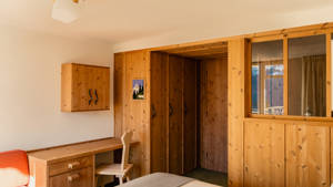 A wooden-finished wardrobe stands in the bedroom, placed near a table with a chair.
