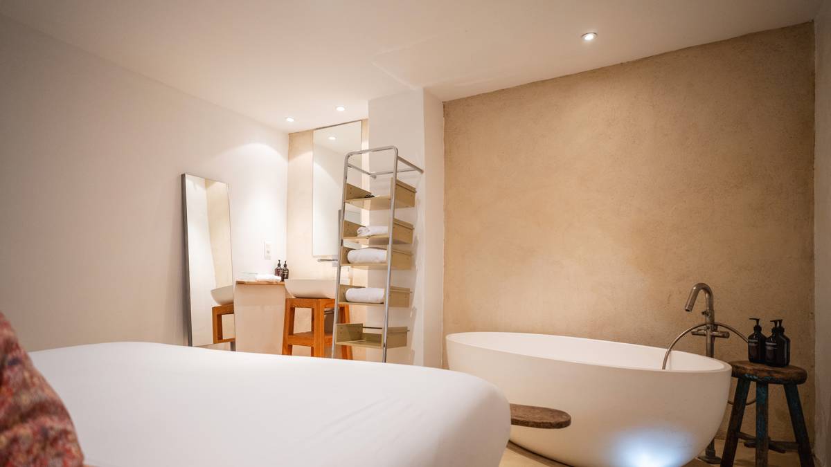 An organized bedroom features a cozy bed, a right-side bathtub, and a chair by mirror.