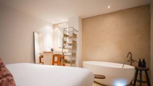 An organized bedroom features a cozy bed, a right-side bathtub, and a chair by mirror.