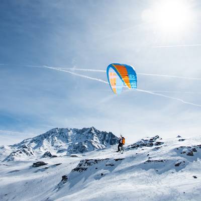 A tandem paragliding pair launching a blue and orange canopy from a snowy mountain ridge under a bright, sunny sky at Val Thorens.