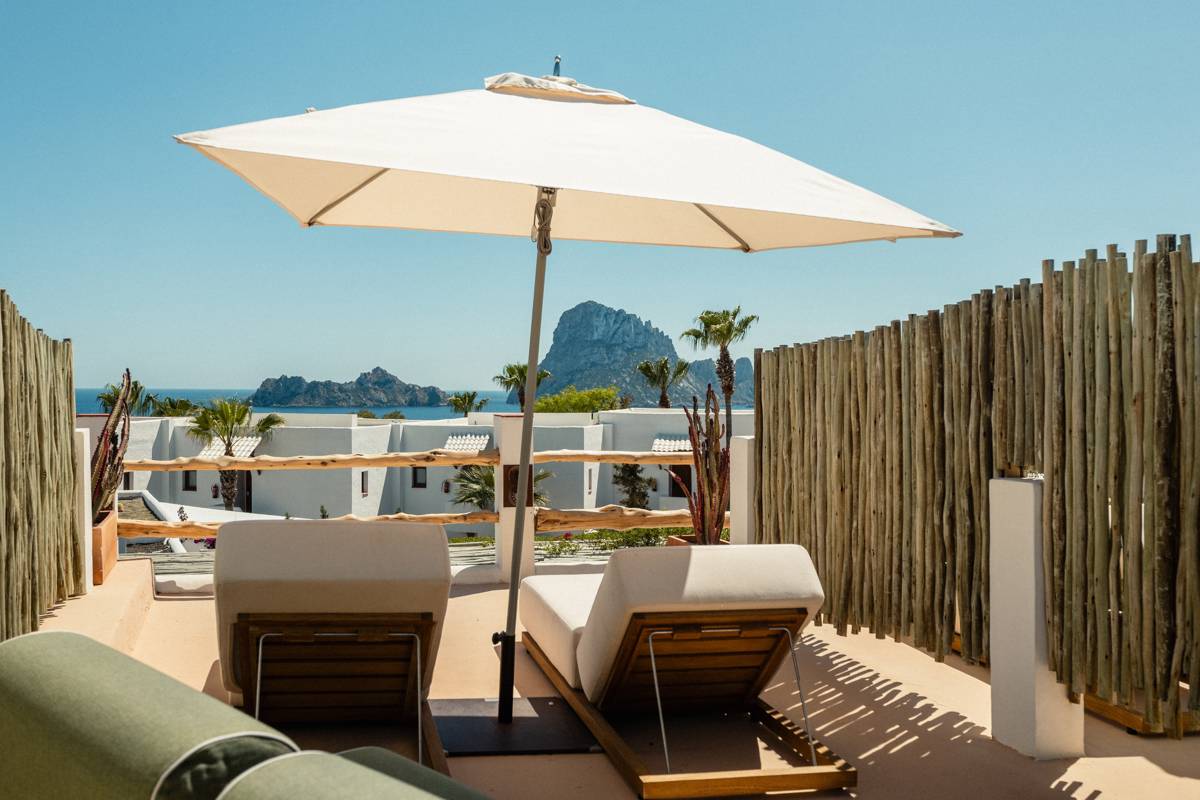 Terrasse ensoleillée avec transats sous un parasol blanc, vue sur mer bleue et îles rocheuses.