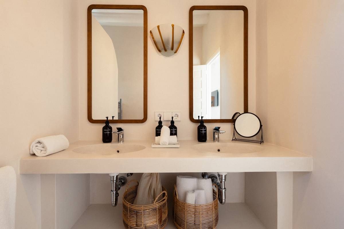 Salle de bain moderne avec deux lavabos, deux miroirs encadrés de bois, des paniers en osier, des distributeurs de savon noirs et une décoration minimaliste dans des tons neutres.
